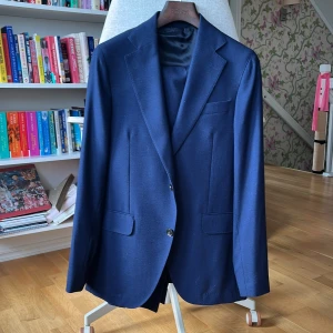 Marinblå kostym från Oscar Jacobson - Snygg marinblå kostym från Oscar Jacobson med klassisk kavaj och matchande byxor. Kavajen har två knappar, fickor med lock och smal krage. Byxorna är raka med pressveck och hällor för bälte. Perfekt för dig som vill ha en stilren och tidlös look. Köptes för ca 5000kr och säljer vidare åt min morbror för betydligt billigare!