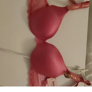 Rosa bh från Juicy Couture ny - Supersnygg rosa bh från Juicy Couture med glansig finish och spetsdetaljer på sidorna. Axelbanden har guldtext med loggan och bh:n har en feminin och trendig look. Perfekt för dig som gillar färg och vill ha något extra i underklädeslådan.