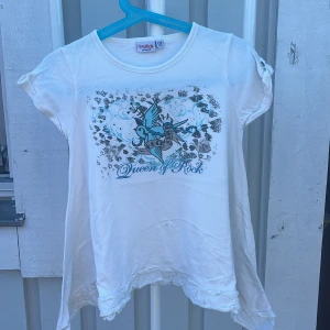 Vit topp Queen of Rock WeKids - Vit topp från WeKids by KappAhl med coolt tryck av en blå fågel, blommor och texten 'Queen of Rock' framtill. Toppen har korta ärmar med knappdetalj och volangkant nertill. Perfekt för dig som gillar rockig stil och snygga detaljer.