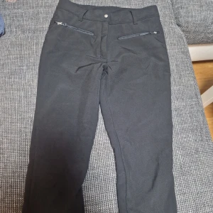 Svarta täckbyxor från Icepeak, strl 36 - Svarta täckbyxor från Icepeak med fleecefoder, dragkedjor vid fickorna och elastiska bensnören. Perfekta för vintern med skön passform och praktiska detaljer som hällor under foten. Snygg och funktionell design för kalla dagar.