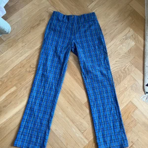 Blå rutiga kostymbyxor - Snygga blå kostymbyxor med rutigt mönster i svart och gult. Byxorna har raka ben, hällor för bälte och två fickor fram. Perfekta för dig som vill sticka ut med färg och mönster. Materialet känns som bomull eller polyester.