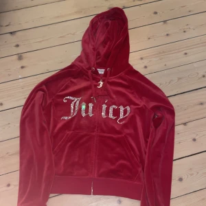 Röd velourjacka Juicy Couture XL - Röd velourjacka från Juicy Couture i storlek XL. Klassisk hoodie med dragkedja, huva och fickor framtill. Stora glittriga Juicy-bokstäver på bröstet och snygga detaljer som J-berlock på dragkedjan. Perfekt för en chill och trendig look.