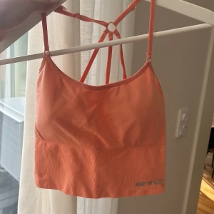 Orange linnetopp från Better Bodies - Snygg orange linnetopp från Better Bodies med tunna axelband och cool ryggdesign med ringdetalj. Perfekt för träning eller när du vill ha en sportig look. Mjuk och stretchig syntetblandning som sitter skönt på kroppen. Liten i storleken