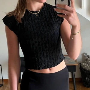 Svart croppad topp med struktur - Säljer en svart croppad topp med snygg struktur och lätt vågiga kanter från Zara i nyskick. Toppen har korta ärmar och en enkel, stilren look som passar perfekt till höga jeans eller kjol. 