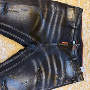 Svarta Dsquared2 jeans med slitningar - Snygga svarta jeans från Dsquared2 med slitna detaljer och vita färgstänk. Jeansen har en smal passform, klassiska fem fickor och en stor vit Dsquared2-patch bak. Materialet är stretchigt denim för extra komfort.