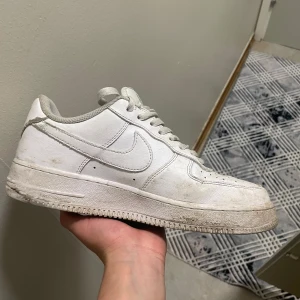 Vita Nike Air Force 1 sneakers - Klassiska vita Nike Air Force 1 sneakers med snörning och rund tå. Skorna har en platt sula och är tillverkade i syntetmaterial och skinn. Perfekta för en stilren och sportig look.