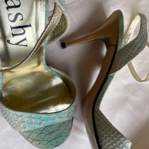 Högklackade exklusiva skor från Lashy. Stl 39 - Säljer ett par supercoola mintgröna skor/sandaletter från Lashy av svenska designern Linda Hammar. Mycket hög kvalité. Tillverkade i portugal. Kalvskinn som liknar mintgrön krokodil! Skorna har öppen tå och hög klack i guld, samt spänne vid ankeln. Sparsamt använda. 