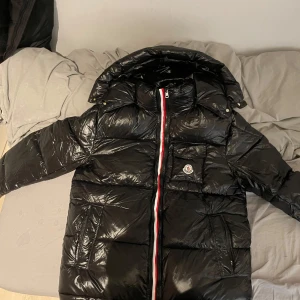 Moncler jacka + NFC - Säljer en snygg svart pufferjacka från Moncler med en dragkedja i rött, vitt och blått. Jackan har en hög krage och en praktisk huva för extra skydd mot väder och vind. Perfekt för kyliga dagar. 
