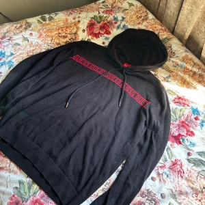 Svart hoodie från Guess med röd logga - Svart hoodie från Guess med röd logga och text på bröstet. Tröjan har huva med dragsko och ribbade muddar vid ärmslut och nederkant. Perfekt för en avslappnad och stilren look.