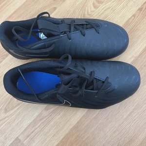 Svarta Nike fotbollsskor med dobbar - Säljer ett par svarta Nike fotbollsskor med vita detaljer och snörning. Skorna har rund tå och platt sula med fasta dobbar för bra grepp på planen. Perfekta för dig som vill ha stilrena och funktionella skor till fotbollsträningen. Skorna är nästintill oanvända 