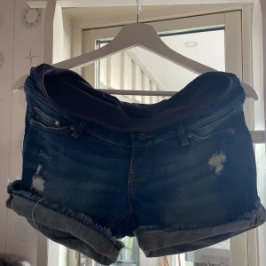 Short jeans från H&M - Mörkblå jeansshorts från H&M med mjuk, stretchig band som är inbyggd i shortsen som ser ut som ett till par shorts under. Shortsen har slitna detaljer, upprullade benslut och klassisk femficksdesign. Perfekta för sommaren och riktigt bekväma.