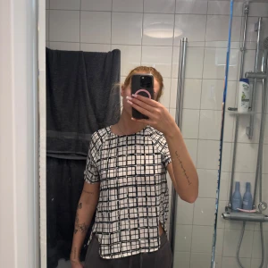 Vit och svart rutig blus från H&M - Säljer en vit blus med svart rutmönster från H&M. Blusen har korta ärmar, rund halsringning och en knappdetalj i nacken. Perfekt för dig som gillar stilrena mönster och enkel design.