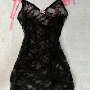 Svart spetsklänning med rosa detaljer - Säljer en svart, genomskinlig spetsklänning med rosa rosett och axelband. Klänningen har v-ringning och är ärmlös, perfekt för dig som gillar feminina detaljer och en lite mer vågad stil.