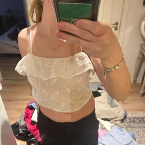 Vit croppad linnetopp med volang från I Love H81 - Säljer en söt vit croppad linnetopp från I Love H81. Toppen har tunna axelband, broderade hålmönster och en volangdetalj framtill. Perfekt för varma dagar och enkel att matcha med jeans eller kjol.