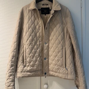 Beige quiltad jacka från Henri Lloyd, strl M - Snygg beige quiltad jacka från Henri Lloyd med klassisk krage och dragkedja framtill. Jackan har fickor med tryckknappar och ett broderat märke på bröstet. Perfekt för övergångssäsonger och stilren look.