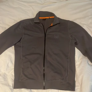 Grå ziptröja från Hugo Boss - Säljer en stilren grå ziptröja från Hugo Boss med hel dragkedja och ståkrage. Tröjan har långa ärmar och diskret logga på bröstet. Perfekt för lager-på-lager och enkel att matcha med det mesta.