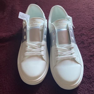 Vita Valentino Garavani sneakers med silverdetaljer - Säljer ett par fräscha vita Valentino Garavani sneakers med breda silverfärgade detaljer och klassisk snörning. Skorna har rund tå och platt sula, samt subtila nitar baktill för en extra touch.