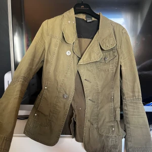 Olivgrön jacka från Diesel - Säljer en olivgrön jacka från Diesel med snygga detaljer som knappar framtill, markerad midja och dekorativa sömmar.