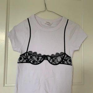 Vit t-shirt med BH-tryck - Vit t-shirt med unik svart spets-bh-tryck. T-shirten har rund halsringning och korta ärmar. Köpt i Australien, finns endast på Glassons. 