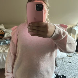 Ljusrosa stickad tröja med volangdetaljer - Säljer en ljusrosa stickad tröja med volangdetaljer på axlarna. Tröjan har lång ärm och är mjuk och bekväm, perfekt för dig som gillar en söt och trendig stil.