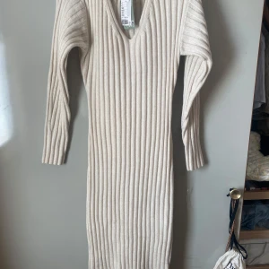 Beige ribbad stickad långklänning med v-ringning - Säljer en beige stickad långklänning med v-ringning och lång ärm. Klänningen har en ribbad struktur som ger en snygg och bekväm passform. Perfekt för dig som gillar stilrena och mysiga plagg. Helt ny med lapp kvar men inte säker på nyspris eftersom att det var en present. Endast testad