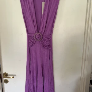 Lila långklänning med djup v-ringning och bältesdetalj - Säljer en lila långklänning med djup v-ringning och markerad midja med dekorativt bälte. Klänningen har ett mjukt, följsamt tyg och är ärmlös, perfekt för sommarens tillfällen.