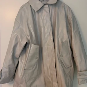 Vit oversized jacka från Weekday - Säljer en vit oversized jacka från Weekday med klassisk krage och stora fickor framtill. Jackan är tillverkad i syntetmaterial med en glansig finish och har långa ärmar samt knappar vid ärmslutet. Se bilder för sprickor som dock inte syns särskilt vid användning.