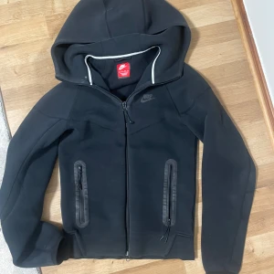 Svart ziphoodie från Nike - Säljer en svart hoodie från Nike med dragkedja och huva. Jackan har två fickor med dragkedja framtill och diskret Nike-logga på bröstet. Perfekt för en sportig och avslappnad stil.