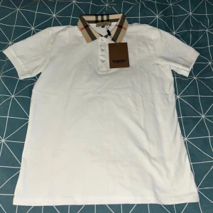 Vit pikétröja med mönstrad krage från Burberry - Säljer en äkta klassisk vit pikétröja från Burberry med deras ikoniska rutiga krage i beige, svart och rött. Tröjan har korta ärmar och knappar framtill. Perfekt för en stilren och trendig look. Kom jättegärna med övriga frågor. Kvitto finns!!!!