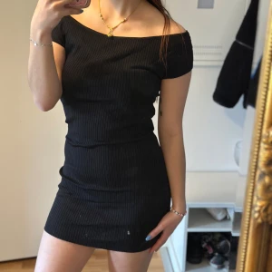 Svart ribbad offshoulder klänning - Säljer en svart, tight ribbad klänning med offshoulder-modell. Klänningen är kort och har en smickrande passform som framhäver figuren. Perfekt för dig som gillar en stilren och enkel look. Aldrig använd