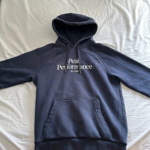 Mörkblå hoodie från Peak Performance - Säljer en mörkblå hoodie från Peak Performance med broderad logga på bröstet. Tröjan har huva med dragsko och en stor magficka framtill. Perfekt för en avslappnad stil och enkel att matcha med jeans eller träningsbyxor.