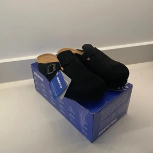 Svarta Birkenstock Boston tofflor - Svarta Birkenstock Boston tofflor i mocka med justerbart spänne och klassisk korksula. Skorna har en rund tå och platt sula, perfekt för en avslappnad och stilren look. Levereras i originalkartong.
