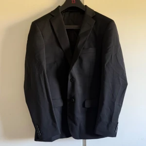 Svart kostym från Dressmann - Säljer en klassisk svart kostym från Dressmann i storlek 48, slim fit. Kavajen har två knappar framtill, fickor med lock och knappar vid ärmslut. Byxorna har pressveck och stängs med knapp och dragkedja. Perfekt för formella tillfällen.