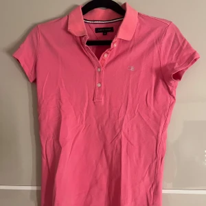 Rosa pikétröja från Tommy Hilfiger - Säljer en klassisk rosa pikétröja från Tommy Hilfiger med korta ärmar och krage. Tröjan har knappar framtill och en liten broderad logga på bröstet. Perfekt för en avslappnad och stilren look.