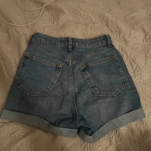 Blå högmidjade jeansshorts - Säljer ett par klassiska blå jeansshorts med hög midja och uppvikta benslut. Shortsen har två bakfickor och två framfickor samt knapp- och dragkedjestängning. Perfekta för sommaren!