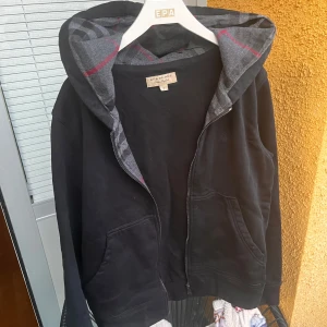 Svart hoodie från Burberry med dragkedja - Svart hoodie från Burberry med klassiskt rutigt foder i huvan och broderad logga på bröstet. Tröjan har dragkedja framtill och två fickor. Perfekt för en stilren och bekväm look. 