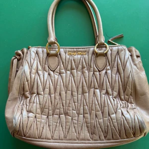 Beige quiltad handväska från Miu Miu - Säljer en beige Matelasse handväska från Miu Miu med guldfärgade metalldetaljer samt löstagbar rem. En liten defekt, se bild. Väl använd. Väskan har dragkedja upptill och flera fack inuti för smart förvaring. Perfekt för dig som vill ha en lyxig och stilren accessoar.