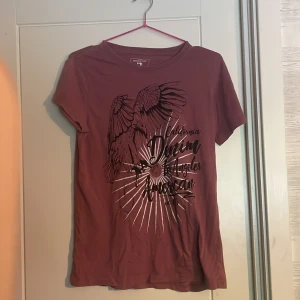 Rostbrun t-shirt med örntryck från Denim Factory - Säljer en rostbrun t-shirt från Denim Factory med ett stort svart tryck av en örn och texten 'California Denim Los Angeles American'. T-shirten har rund hals och korta ärmar. Perfekt för dig som gillar coola tryck och streetstyle.