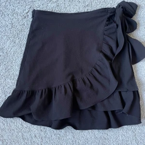 Svart omlottkjol med volang från Vero Moda - Supersnygg svart omlottkjol från Vero Moda med volangdetaljer och knytband i midjan. Kjolen är kort och har en feminin och trendig look som passar perfekt till många olika stilar. Kjolen har små knottror i tyget, men det är inget som förstör helhetsintrycket🌸🌸🌸