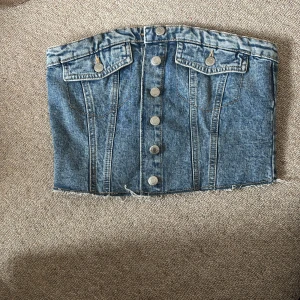 Jeans tubtopp med knappar - Tubtopp i blå denim med knappar framtill. Toppen har en croppad passform och elastisk smock baktill så den passar fler former. Den är använd 1 gång och är i nyskick.