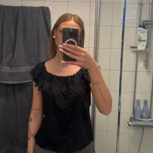 Svart offshoulder topp med spets från Miss Cocoa - Säljer en svart offshoulder topp från Miss Cocoa med dekorativ spets längs överkanten. Toppen har en lös passform och är perfekt för dig som gillar en stilren men ändå detaljrik look.