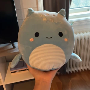 Blå Squishmallows drak-gosedjur - Supermjuk och rund blå drak-gosedjur från Squishmallows med vita detaljer på magen och små silverprickar på huvudet. Har små vingar, horn och taggar längs ryggen. Perfekt att krama eller ha som dekoration.