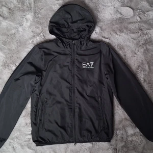 Svart vindjacka från EA7 Emporio Armani - Snygg svart vindjacka med huva från EA7 Emporio Armani. Jackan har dragkedja framtill, två fickor med dragkedja och logga på bröstet. Lätt och smidig modell med meshfoder på insidan. Använd vid 3 tillfällen.  Stl XS/170