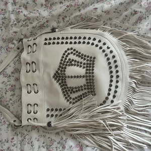 Vit axelväska med nitar och fransar - Säljer en vit axelväska med coola nitar i mönster på framsidan och långa fransar längs kanterna. Väskan har bred axelrem och dekorativa snörningar. Mycket plats skriv för fler frågor mm