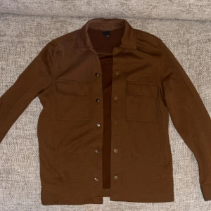 Brun overshirt från H&M - Säljer en brun overshirt från H&M med två bröstfickor och knappar framtill. Skjortan har klassisk krage och är långärmad, perfekt att styla lager på lager. Materialet känns mjukt och passar bra till alla årstider.