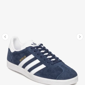 Adidas Gazelle blå sneakers - Säljer ett par klassiska Adidas Gazelle sneakers i mörkblå mocka med vita detaljer och snörning. Skorna har de ikoniska tre ränderna på sidan och en vit sula. Perfekta för dig som gillar stilrena och sportiga skor.