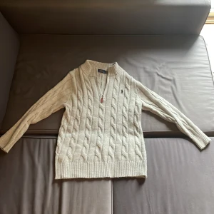 Grå kabelstickad tröja från Polo Ralph Lauren - Säljer en grå  kabelstickad tröja från Polo Ralph Lauren med halv dragkedja och liten broderad logga på bröstet.