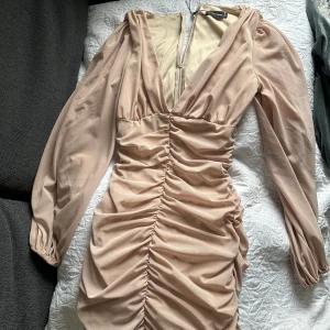 Beige klänning från PLT - Säljer en beige klänning från pretty little thing, med  v-ringning och långa ärmar. Klänningen har rynkade detaljer längs hela framsidan och en dragkedja i ryggen. 