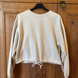 Beige sweatshirt med dragsko från Gina Tricot - En beige sweatshirt från Gina Tricot med rund halsringning och dragsko i nederkanten. Tröjan har långa ärmar och en avslappnad passform, perfekt för en chill och stilren look. Storlek S.