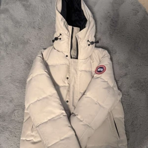 Vit dunjacka från Canada Goose - Säljer en snygg vit dunjacka från Canada Goose med huva och svart innerfoder. Jackan har dragkedja, knappar och justerbar snörning vid huvan. Perfekt för kalla dagar och har den klassiska Canada Goose-loggan på bröstet.